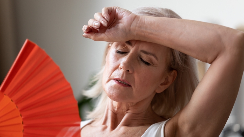 A woman uses a hand fan and wipes her forehead, showing signs of a hot flush during menopause
