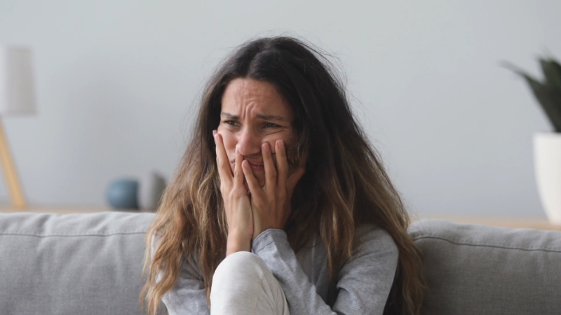 A woman sitting on a couch, visibly distressed and holding her face in her hands