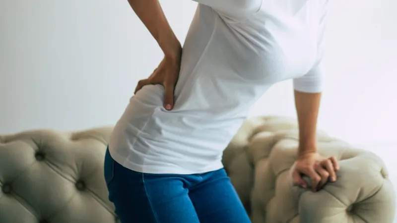 A woman holds her lower back in pain while standing up, showing signs of chronic back discomfort