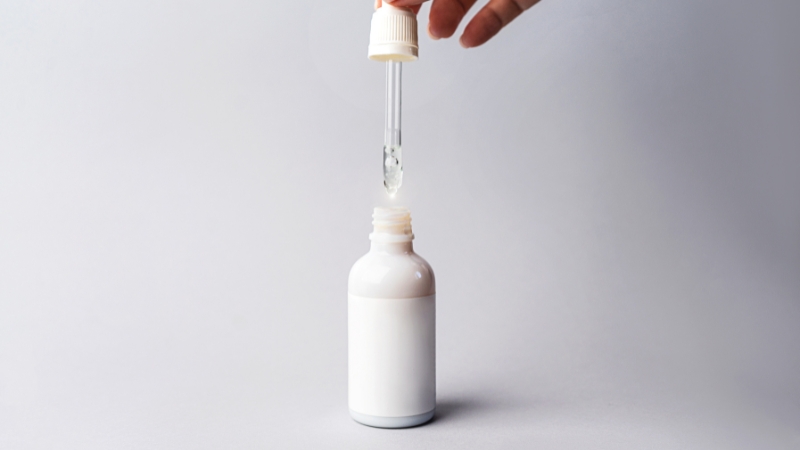 A white dropper bottle with clear retinol serum being dispensed from a glass pipette