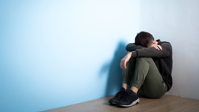 A man sits alone in a corner with his head down and arms over his knees, showing signs of emotional distress