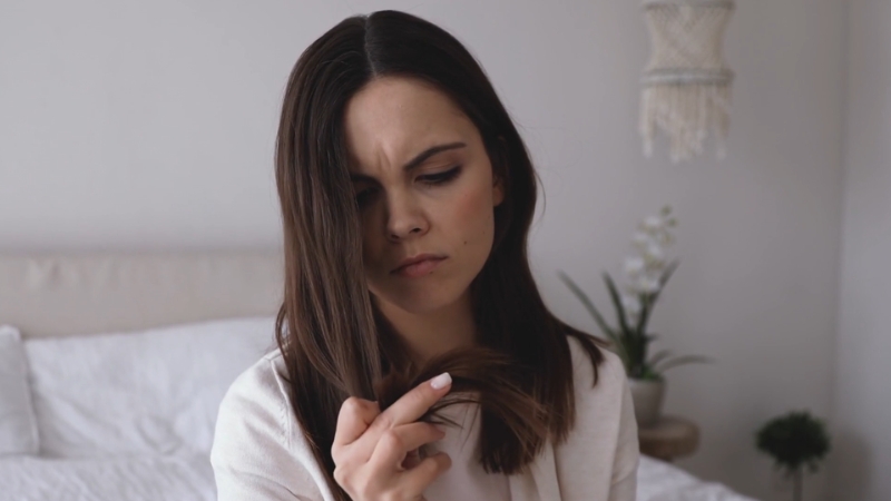 Woman examining the ends of her hair with concern, showing signs of hair loss possibly linked to poor nutrition