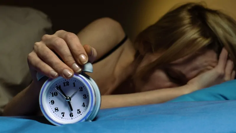 Woman struggling to wake up while holding an alarm clock, showing effects of poor sleep on health