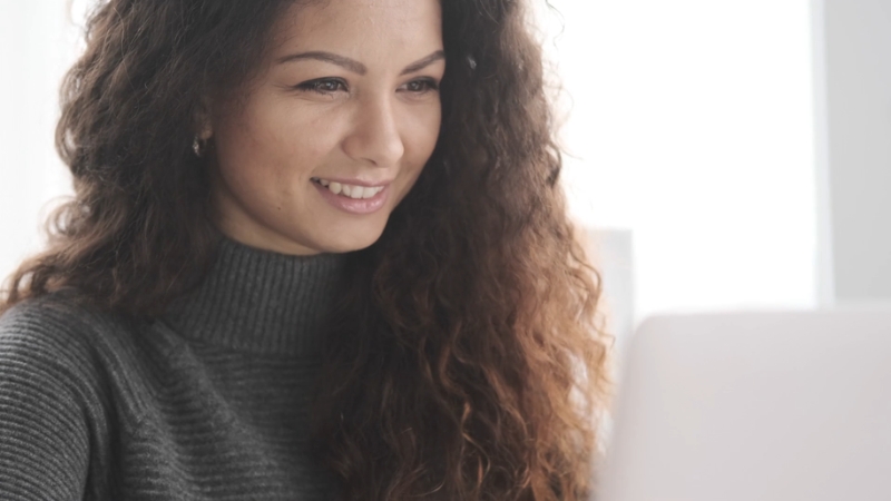 A woman with long curly hair smiles while looking at her laptop