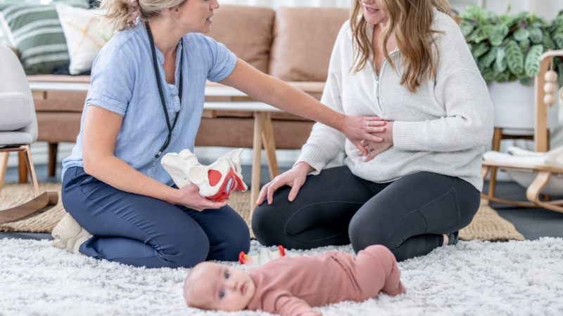 A healthcare worker checks a new mom's belly as they sit on the floor, showing gaps in routine postpartum care