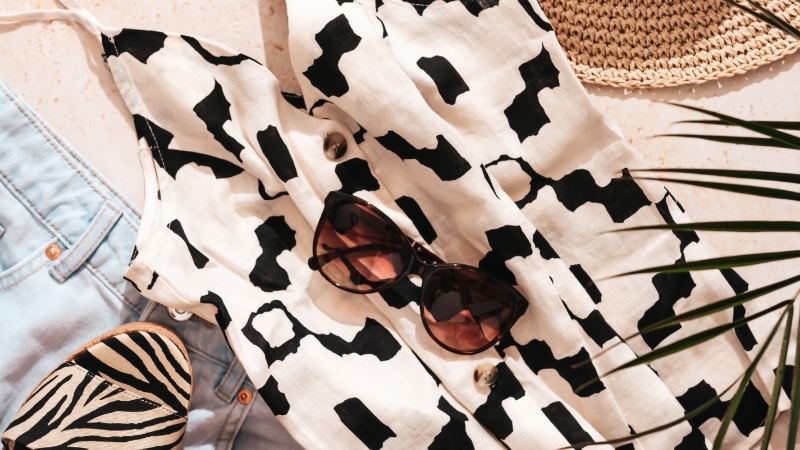 A patterned summer dress, sunglasses, and zebra sandals are laid out with denim and a straw hat for a bold, coordinated summer look