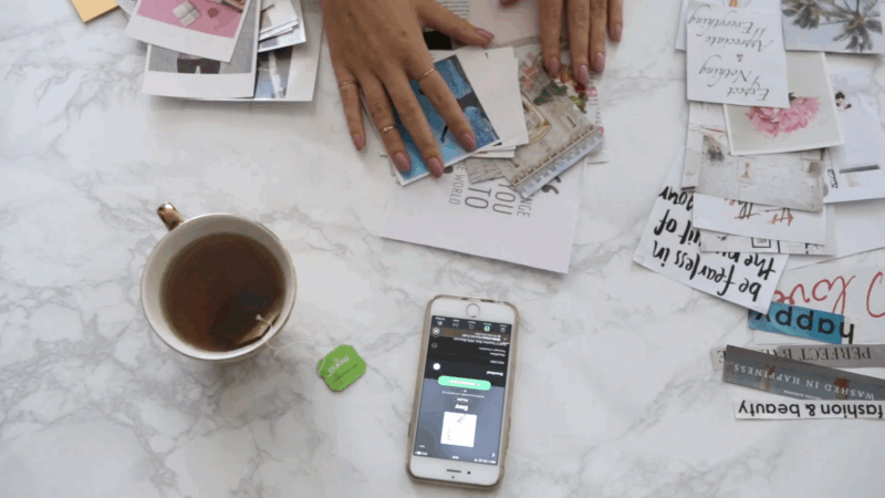 Hands sorting through printed photos and quotes on a table with tea and a phone nearby