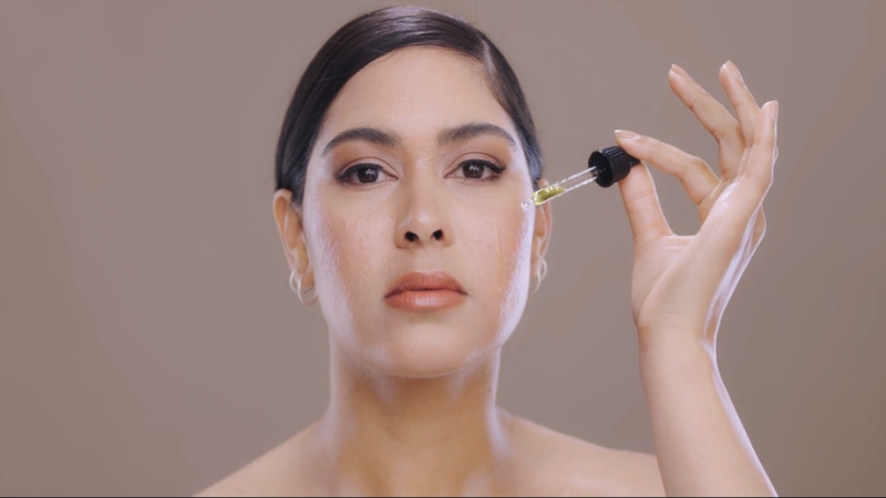 A woman applies retinol serum to her face with a dropper against a neutral background