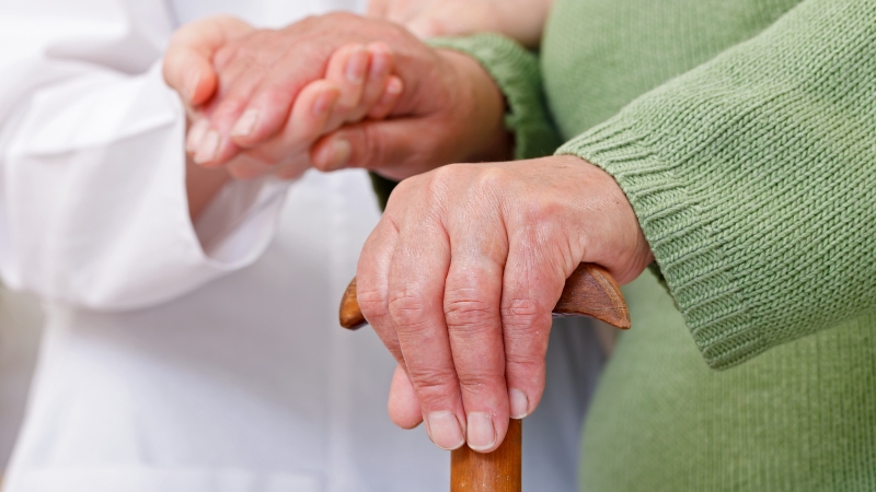 A woman in a green sweater holds a wooden cane while a caregiver gently supports her hand