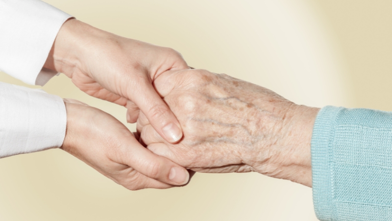 A caregiver gently holds an elderly woman's hand, reflecting the quiet struggles women face in nursing homes