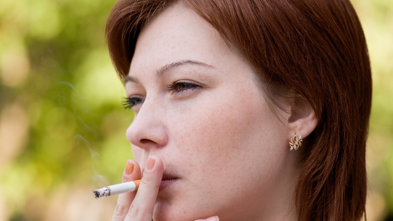 A woman with short auburn hair smoking a cigarette outdoors, showing a habit linked to hair loss