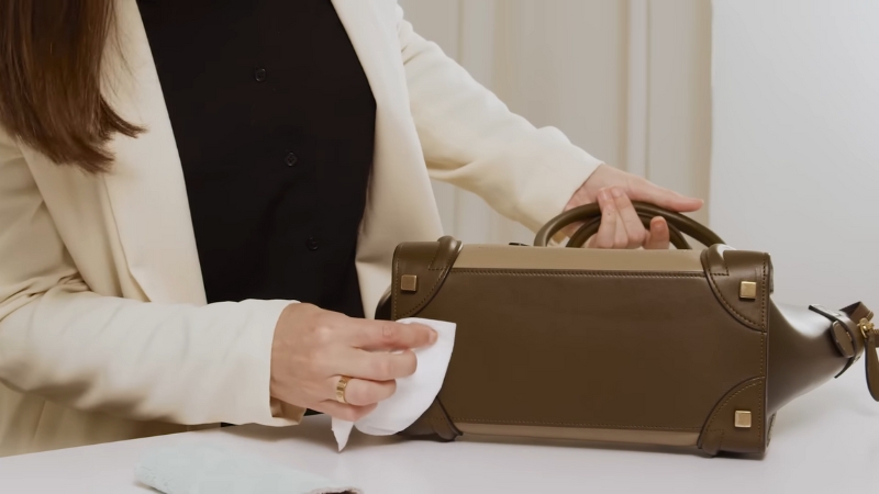 Person cleaning a structured olive green leather handbag using a white cloth
