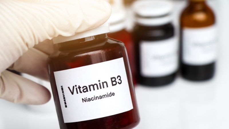 A gloved hand holds a dark glass bottle labeled Vitamin B3 Niacinamide
