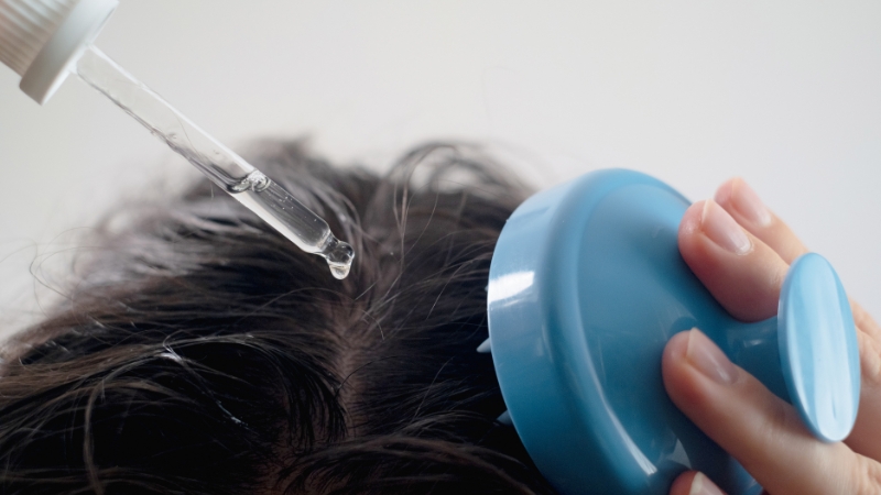 A person applying a liquid scalp treatment with a dropper while using a blue scalp massager