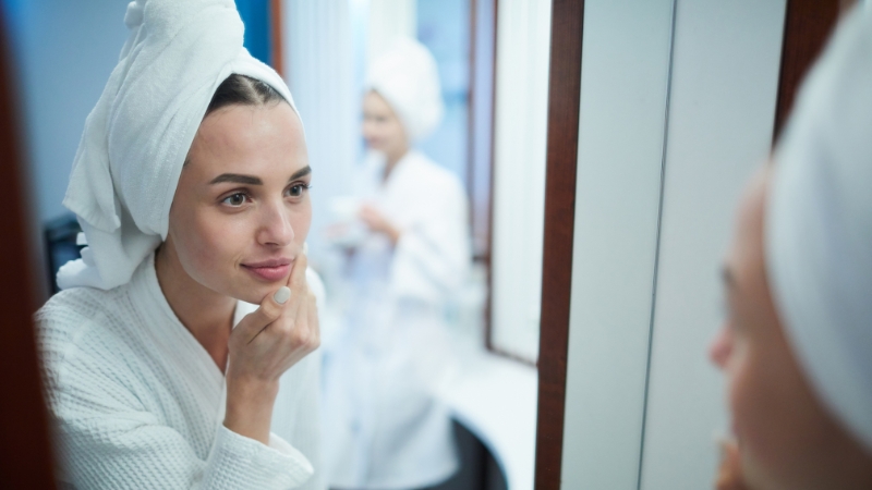 A woman in a bathrobe applies skincare in front of a mirror while another family member gets ready in the background