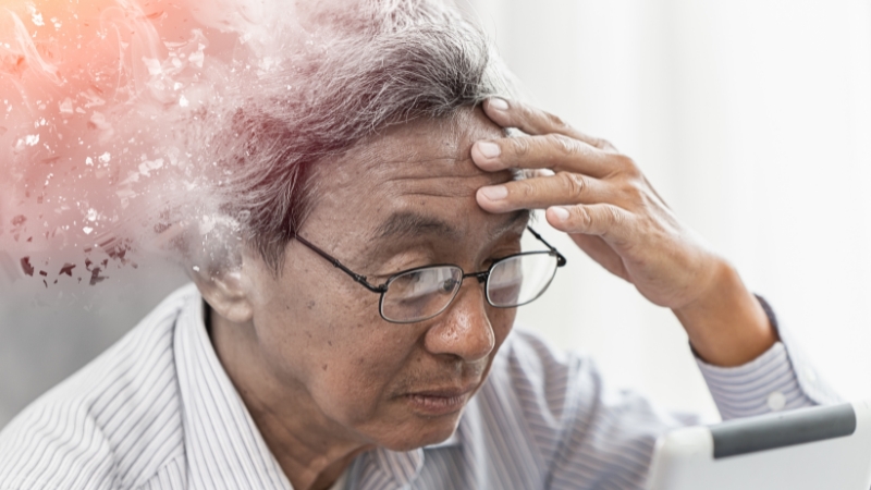 An elderly man with glasses holds his forehead while looking at a tablet