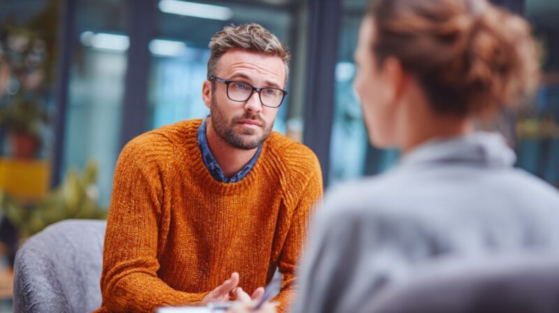 A man in an orange sweater and glasses talking to a woman in a gray top, seated in a modern office setting