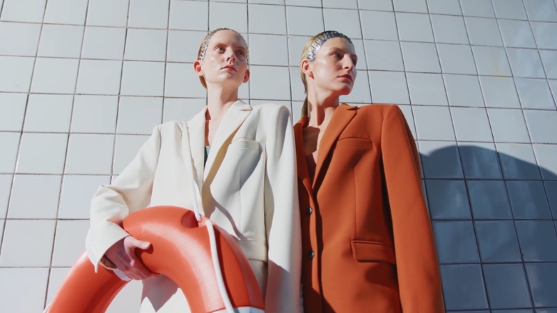 Two models pose in structured suits against a tiled wall during a fashion shoot