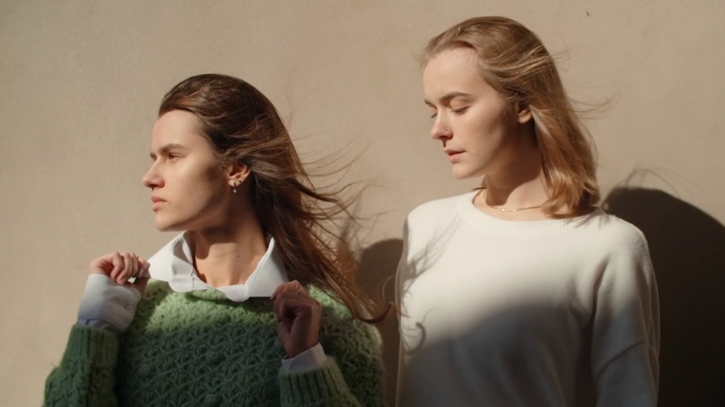 Two models stand in soft sunlight against a plain wall during a casual outdoor shoot