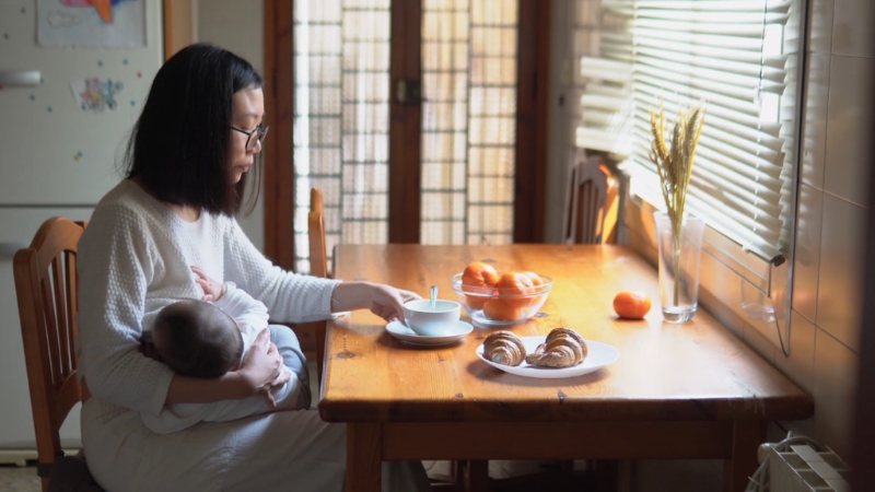 Mother breastfeeding at the table with tea, fruit, and pastries