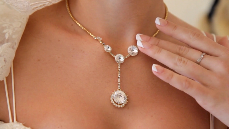 A woman wearing a gold necklace with large crystal stones touches it gently for a close-up shot